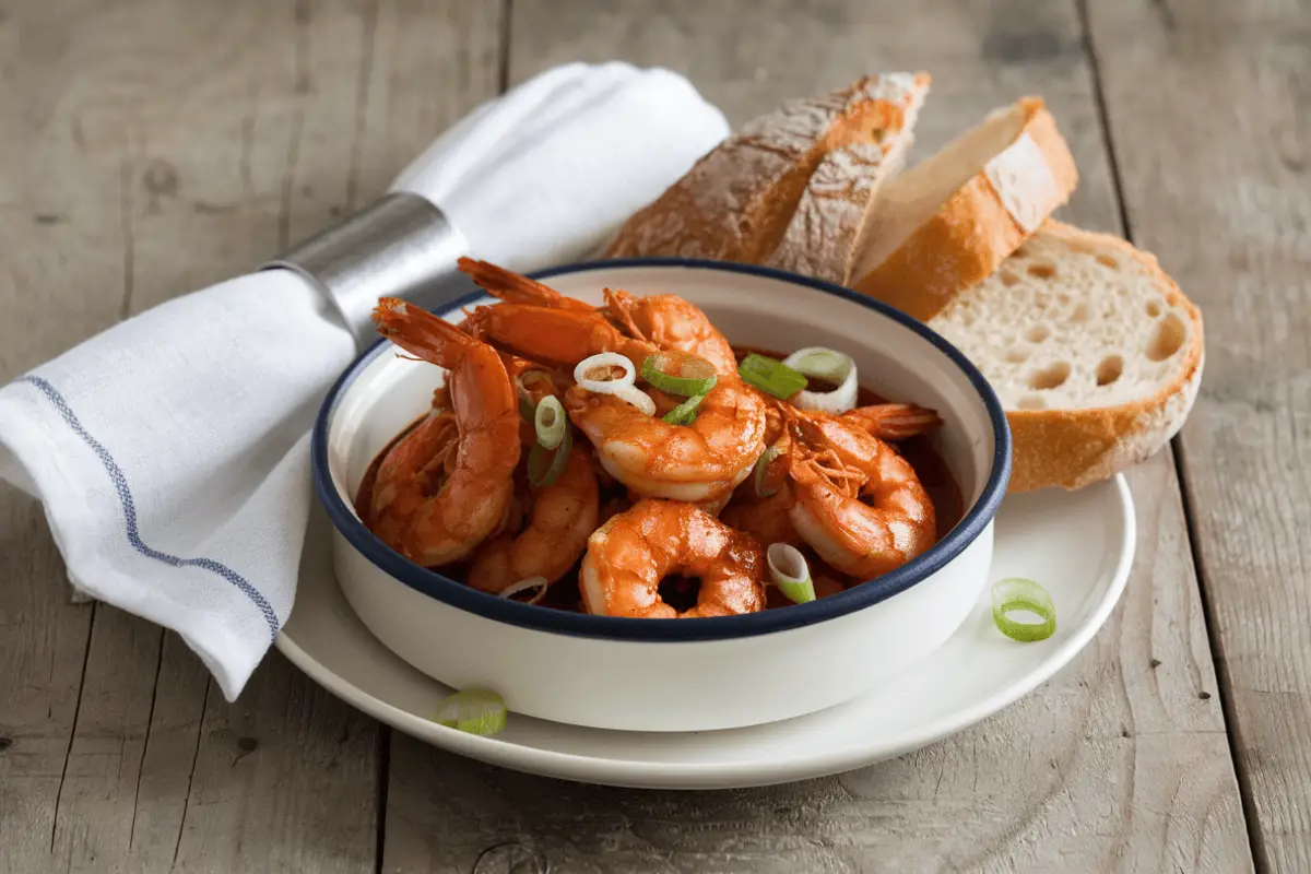 Plated barbecue shrimp recipe with rich red sauce, garnished with green onions and served with crusty bread.